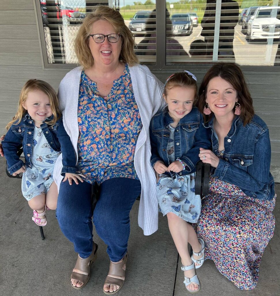 A picture of one of Elasticity's co-workers, Miranda Oschner, kneeling next to a bench that sits her two daughters and her mom, Linda. Miranda and her two kids have jean jackets and dresses on, and her mom is in a sweater, shirt and jeans. All four of smiling.