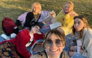 A group of five female friends are sitting on a blanket in the park with books, backpacks and snacks. One is holding the camera for a picture up close while the rest are smiling and making silly faces for the camera. 
